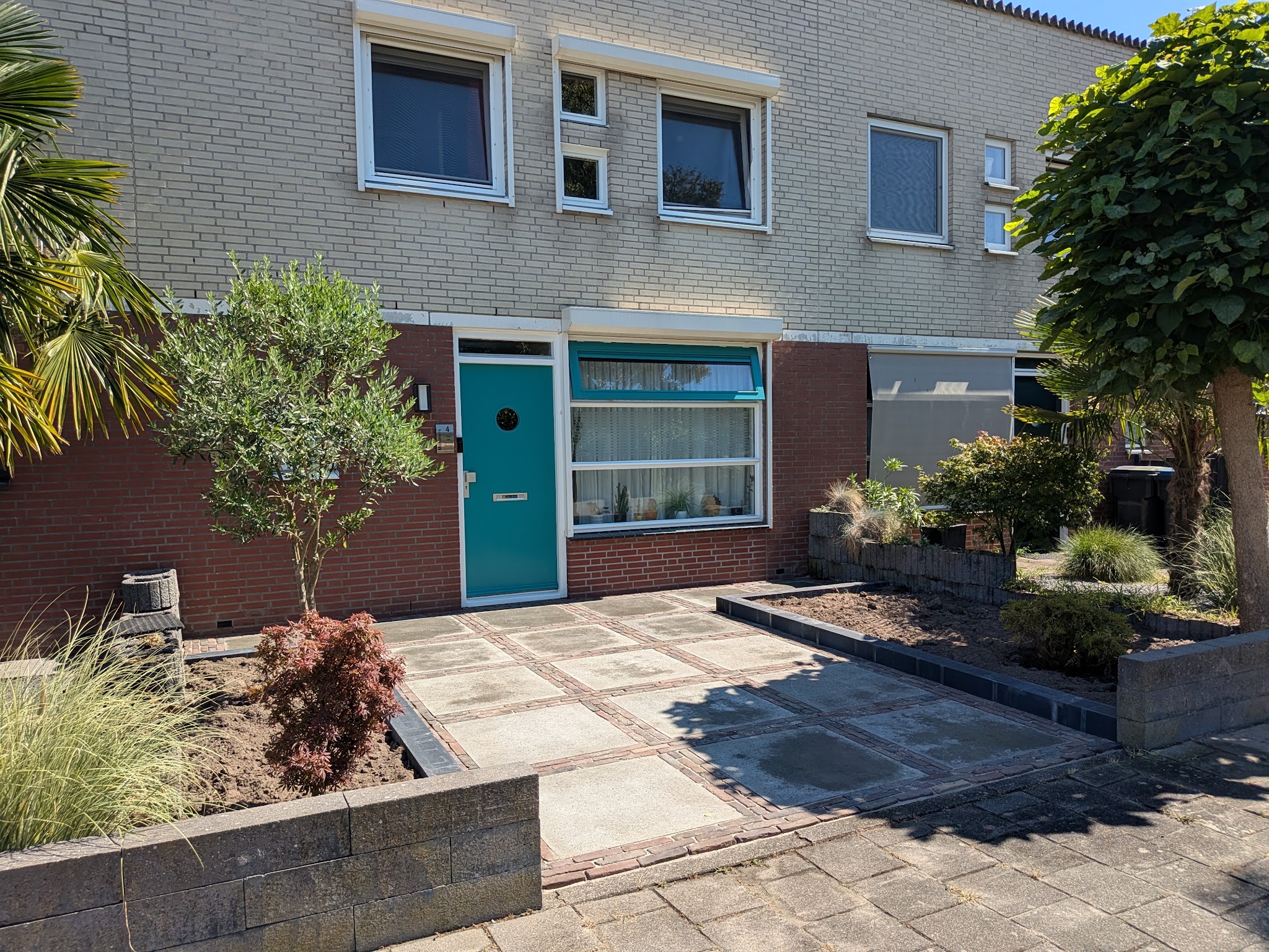Vernieuwde voortuin met grote grijze tegels, banden van oude klinkers en verhoogde plantenbakken