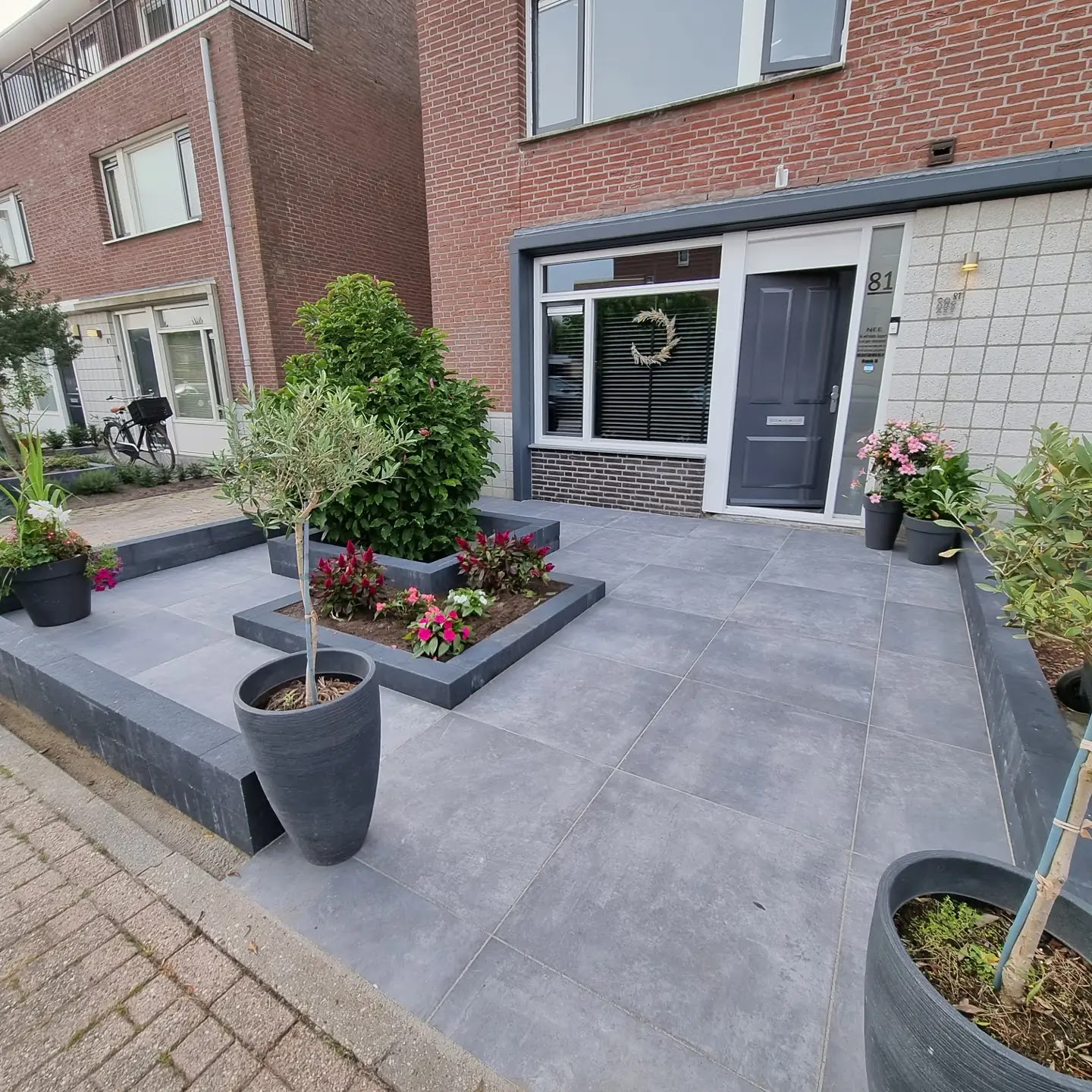 Voortuin met grote antraciet tegels, verhoogde plantenbakken en een olijfboompje