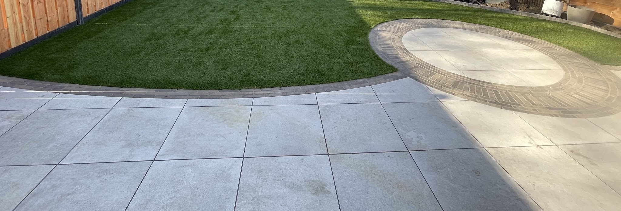 Achtertuin met kunstgras, een golvende rand van klinkers en een rond terras van tegels in een klinkerring